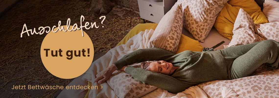 Ausschlafen? Tut gut! – Frau in gemütlicher Kleidung streckt sich lächelnd auf einem Bett mit gemusterter Bettwäsche