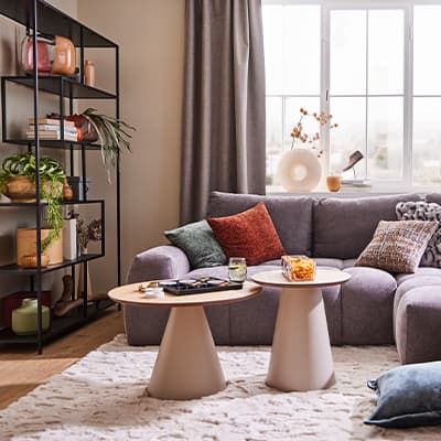 Gemütliches Wohnzimmer mit grauem Sofa, bunten Kissen, zwei runden Couchtischen und Regal mit Pflanzen und Dekorationen