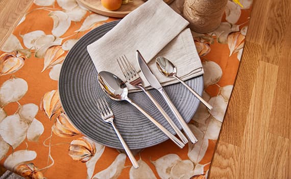 Besteck-Set auf grauem Teller mit beigem Stoffserviette, auf orangem Tischläufer mit Blumendesign, Holzuntergrund