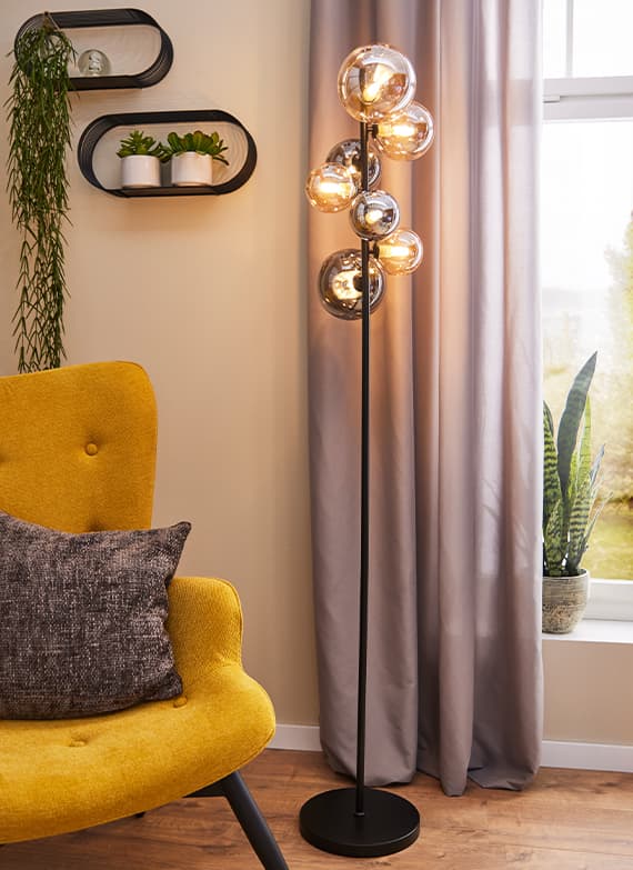 Moderne Stehlampe mit mehreren Leuchtkugeln neben gelbem Sessel mit Kissen, grauer Vorhang im Hintergrund, Pflanzen auf Fensterbank und Wandregal