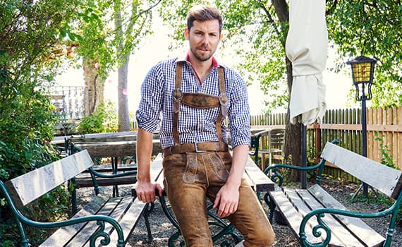 Mann in traditioneller bayerischer Lederhose und kariertem Hemd sitzt auf einer Holzbank in einem Garten mit Bäumen und Zaun.