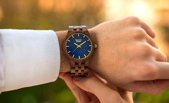 Person trägt Uhr mit blauem Zifferblatt und Holzarmband, im Hintergrund verschwommene Natur und Sonnenlicht