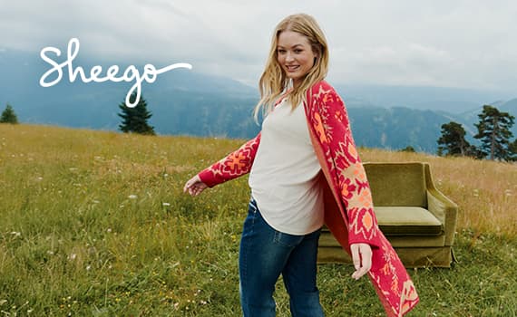 Sheego – Frau in weißem Shirt und rotem Cardigan steht auf einer Wiese, Berge im Hintergrund