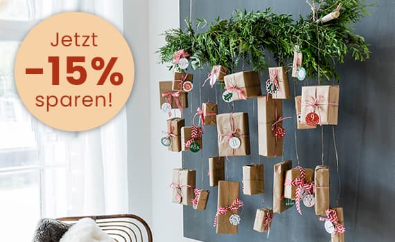 Jetzt -15% sparen! Adventskalender aus braunen Papiertüten mit roten und grünen Schleifen an Wand befestigt