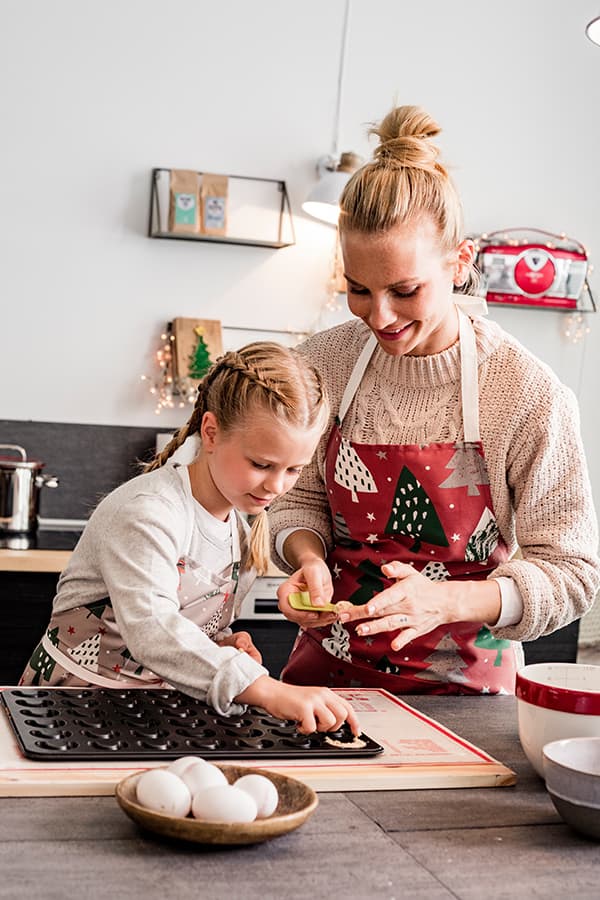 Kind und Erwachsene backen zusammen Plätzchen in der Küche, beide tragen festliche Schürzen mit Weihnachtsmotiven.