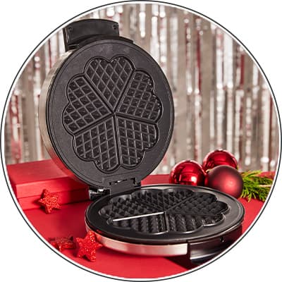 Geöffneter Waffeleisen mit Herzformen, dekoriert mit roten Weihnachtskugeln und Sternen auf rotem Untergrund