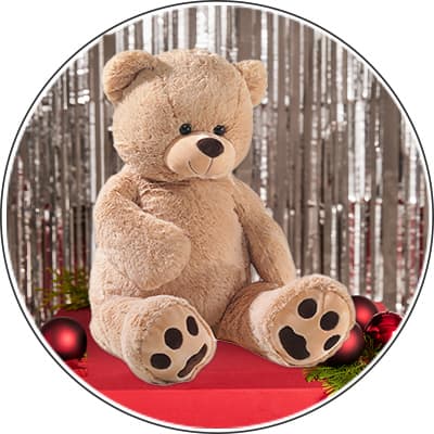 Großer Plüschbär sitzt auf rotem Tisch mit Weihnachtskugeln, im Hintergrund silberne Lametta-Dekoration