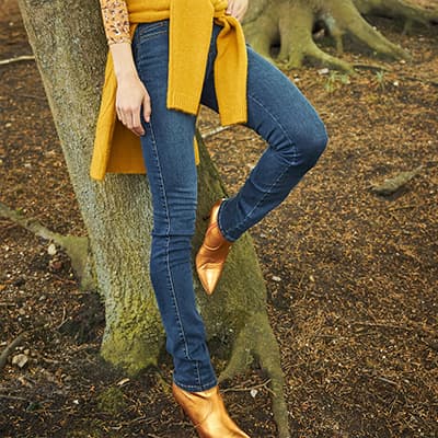 Frau steht im Wald, trägt blaue Jeans und goldene Stiefeletten, gelber Schal um die Taille.