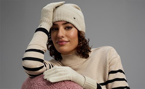 Person in beigem Strickpullover mit schwarzen Streifen, passenden Handschuhen und Mütze, lehnt auf pinkem Plüschkissen