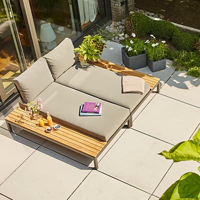 Outdoor-Lounge mit grauem Polster, Buch, Getränken und Pflanzen, umgeben von Garten.