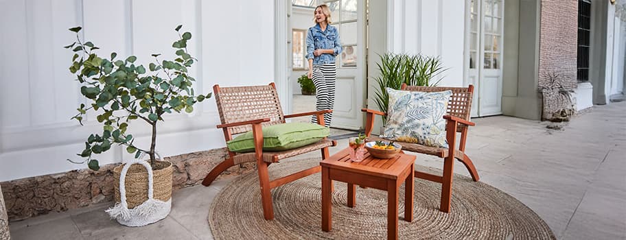 Zwei Holzstühle mit Kissen und ein Tisch auf einer Terrasse, Pflanze im Korb, Person im Hintergrund.
