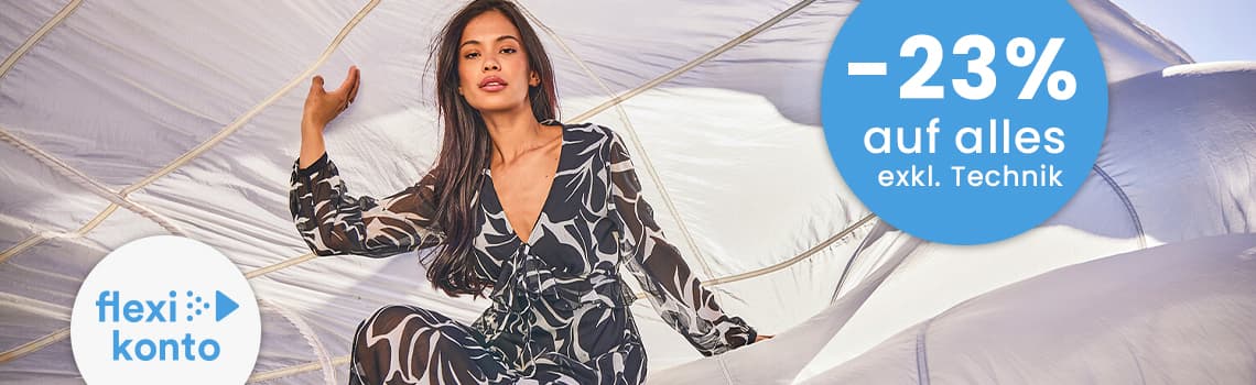 -23 % auf alles exkl. Technik – Frau im gemusterten Jumpsuit sitzt zwischen aufgeblähten weißen Stoffbahnen