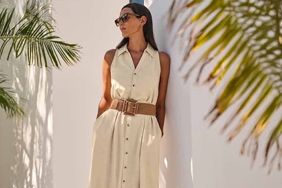 Frau in ärmellosem Hemdkleid mit breitem Gürtel lehnt an weißer Wand im Schatten einer Palme