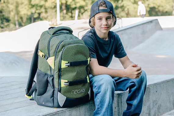 Kind mit Rückwärtskappe sitzt am Skatepark neben großem Schulrucksack mit gelben Details