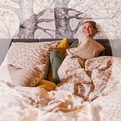 Frau sitzt im Bett zwischen gemusterter Bettwäsche und vielen Kissen und hält lachend ein Kissen im Arm