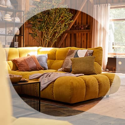 Gelbes Sofa mit vielen Kissen in warmem Wohnzimmer, daneben Couchtisch, Regal und große Pflanze im Hintergrund