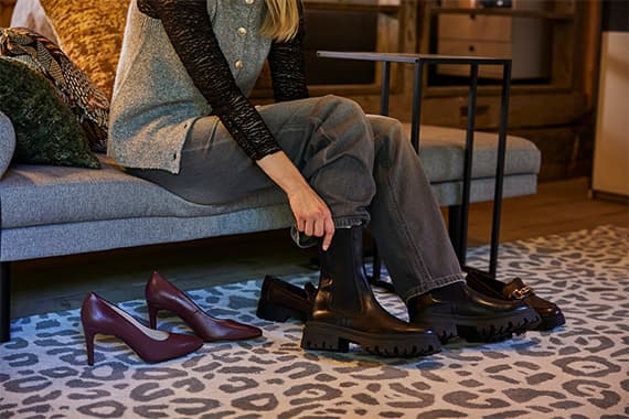 Frau sitzt auf Sofa, zieht klobigen Stiefel an, neben ihr auf dem Teppich stehen elegante Pumps und Loafer